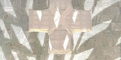 Ein in weißen Stoff gehülltes Kreuz hängt vor einer grauen Wand, Schatten von Palmzweigen überdecken alles.