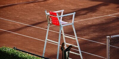 ein Tennisplatz mit rotem Sand und weißen Linien, daneben ein leerer weißer Schiedsrichterstuhl